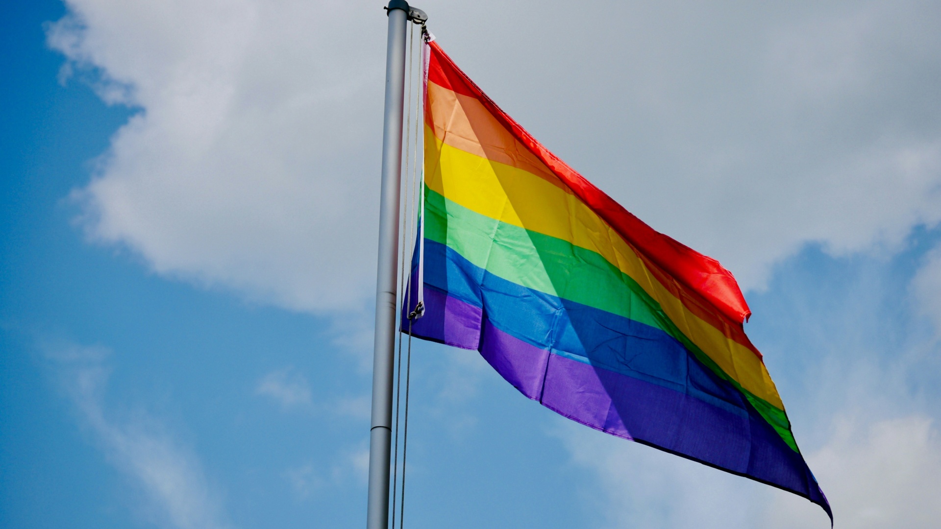 A rainbow-striped Pride flag flies against a blue sky background
